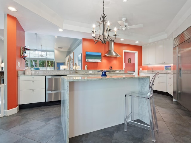 kitchen featuring pendant lighting, white cabinets, wall chimney range hood, built in refrigerator, and a tray ceiling