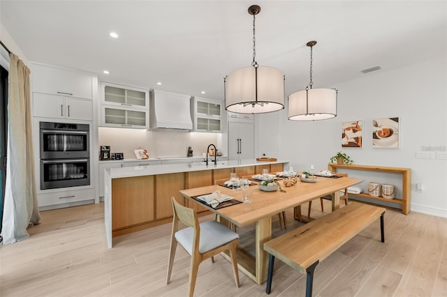 kitchen with custom range hood, stainless steel double oven, decorative light fixtures, white cabinets, and an island with sink