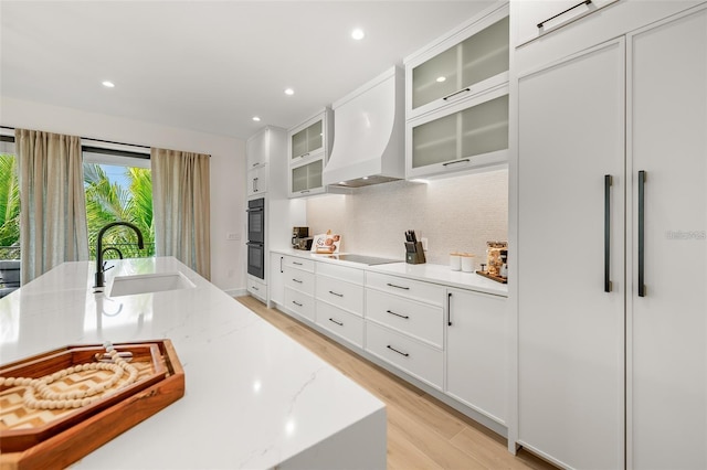 kitchen with light stone countertops, custom range hood, black appliances, sink, and light hardwood / wood-style flooring