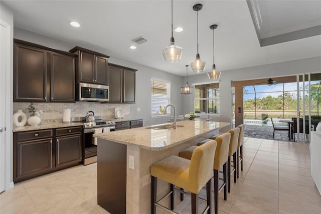 kitchen with light stone countertops, appliances with stainless steel finishes, a kitchen island with sink, sink, and decorative light fixtures