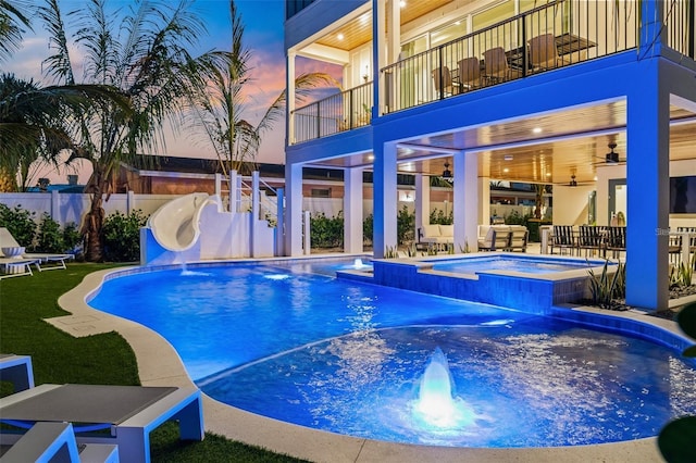 pool at dusk featuring an in ground hot tub, pool water feature, ceiling fan, a patio area, and a water slide