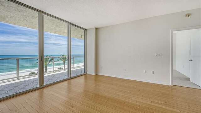 empty room featuring a view of the beach, a water view, light hardwood / wood-style flooring, a textured ceiling, and a wall of windows