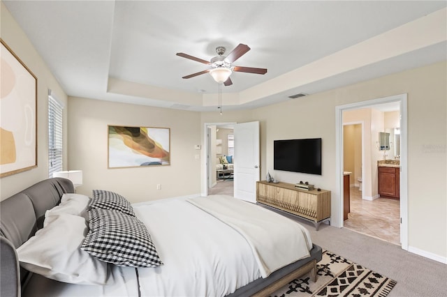 bedroom with ceiling fan, ensuite bathroom, light carpet, and a tray ceiling