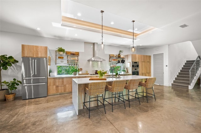 kitchen featuring a large island with sink, a raised ceiling, wall chimney range hood, hanging light fixtures, and stainless steel appliances