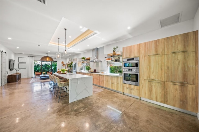 kitchen featuring a breakfast bar, a tray ceiling, a spacious island, wall chimney range hood, and a barn door