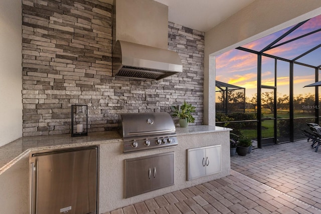 patio terrace at dusk featuring a lanai, area for grilling, and an outdoor kitchen