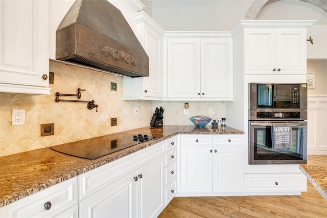 kitchen with custom exhaust hood, black appliances, dark stone countertops, tasteful backsplash, and white cabinetry