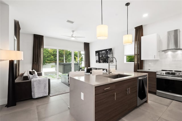 kitchen with stainless steel appliances, sink, wall chimney range hood, decorative light fixtures, and a center island with sink