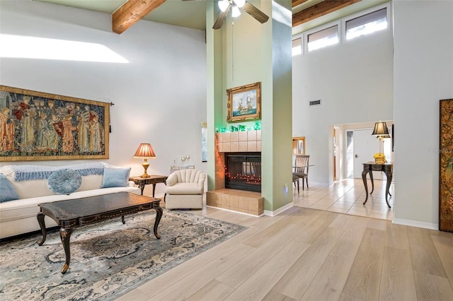 living room featuring ceiling fan, a high ceiling, beamed ceiling, a tiled fireplace, and light wood-type flooring