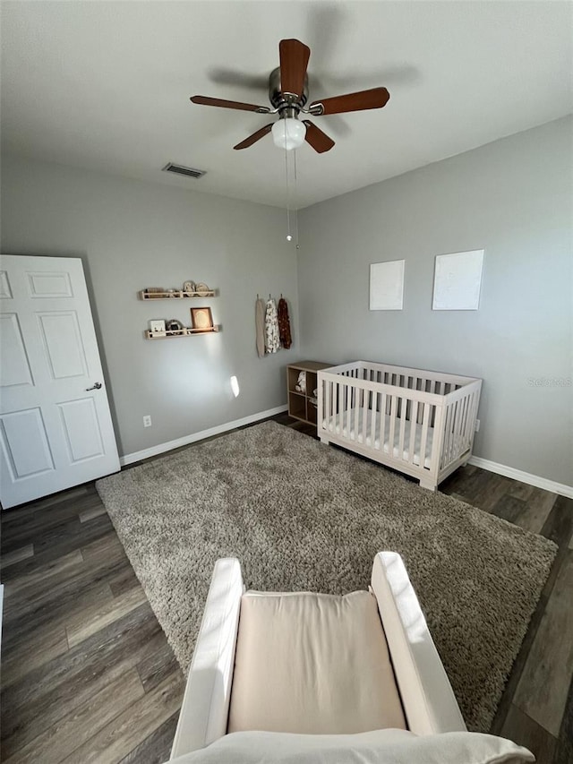 bedroom with a crib, ceiling fan, and dark wood-type flooring