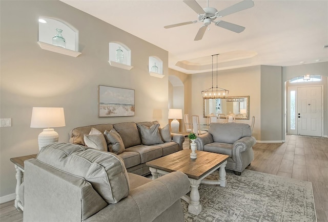 living room with ceiling fan with notable chandelier, light wood-type flooring, and a tray ceiling