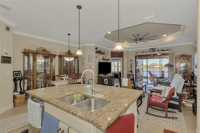 kitchen with a kitchen island with sink, a raised ceiling, sink, light stone countertops, and decorative light fixtures