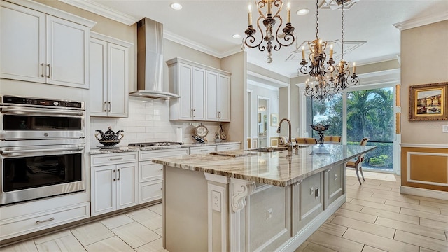 kitchen featuring appliances with stainless steel finishes, a kitchen island with sink, wall chimney range hood, pendant lighting, and white cabinetry