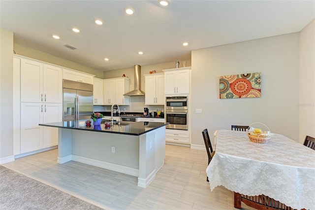 kitchen featuring stainless steel appliances, white cabinetry, a center island with sink, and wall chimney range hood