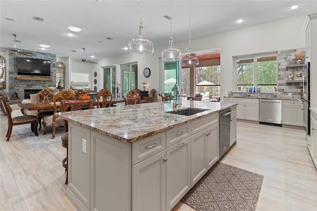 kitchen featuring a kitchen island with sink, ceiling fan, sink, a stone fireplace, and hanging light fixtures