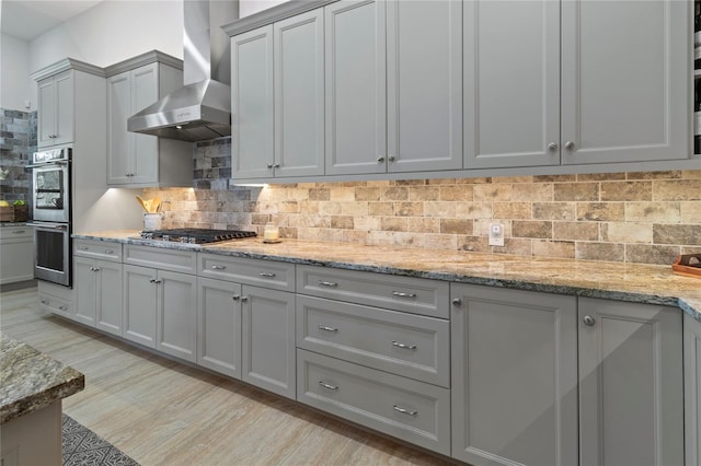 kitchen with gray cabinetry, wall chimney exhaust hood, light stone countertops, appliances with stainless steel finishes, and tasteful backsplash