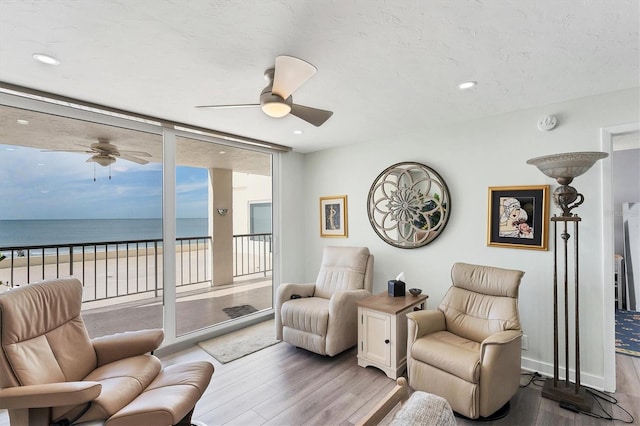 sitting room featuring ceiling fan, a beach view, expansive windows, light hardwood / wood-style floors, and a water view