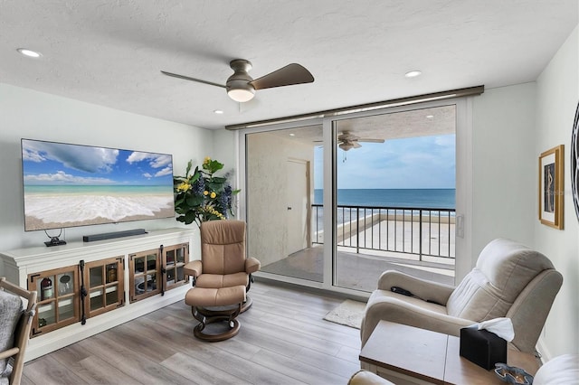 living room with ceiling fan, a water view, a wall of windows, a beach view, and light hardwood / wood-style floors