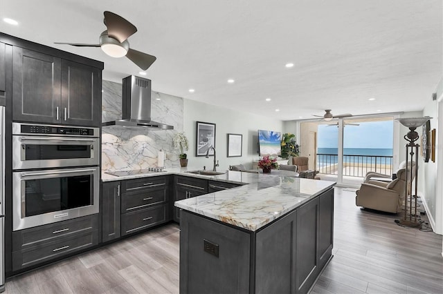 kitchen featuring decorative backsplash, light wood-type flooring, double oven, sink, and wall chimney range hood