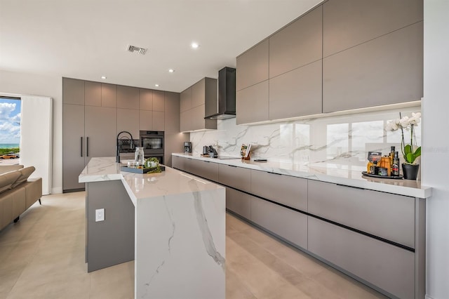 kitchen featuring a kitchen island with sink, wall chimney range hood, gray cabinets, black electric cooktop, and light stone counters