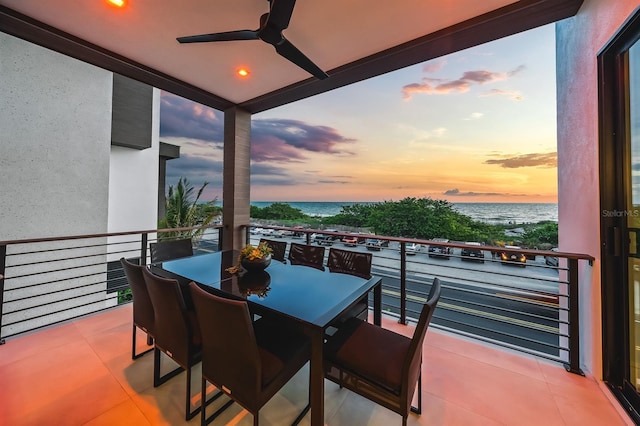 balcony at dusk with a beach view, a water view, and ceiling fan