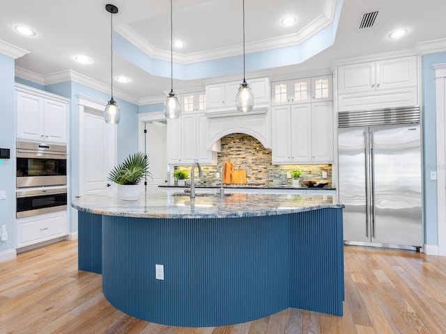 kitchen with appliances with stainless steel finishes, light stone counters, a tray ceiling, a kitchen island with sink, and white cabinetry