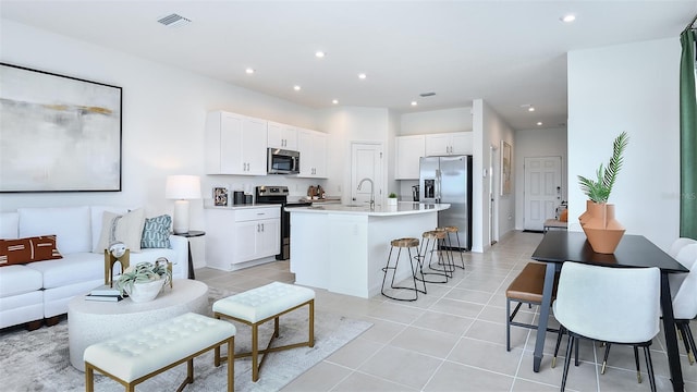 kitchen with a kitchen bar, stainless steel appliances, a kitchen island with sink, light tile patterned floors, and white cabinetry