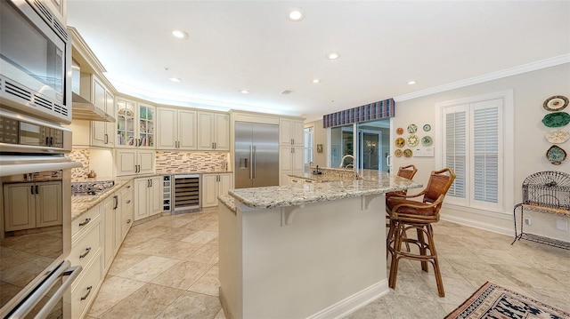 kitchen featuring decorative backsplash, a kitchen bar, a kitchen island with sink, built in appliances, and cream cabinetry