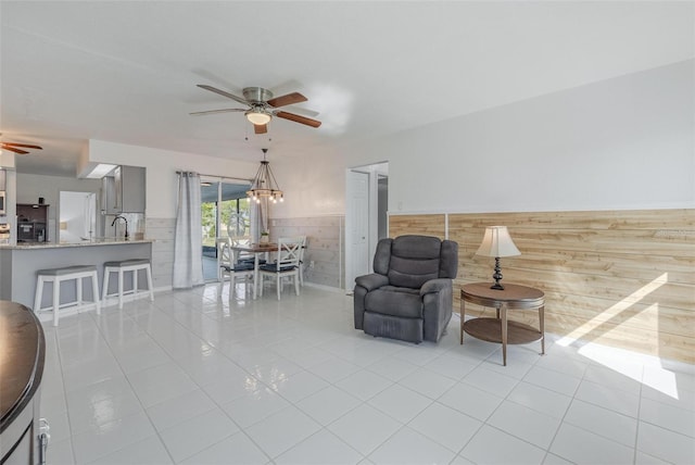 tiled living room with ceiling fan with notable chandelier, wood walls, and sink