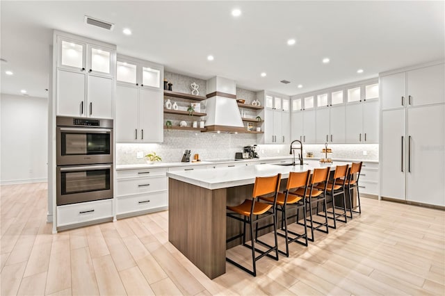 kitchen featuring custom exhaust hood, a breakfast bar, a center island with sink, white cabinetry, and stainless steel double oven