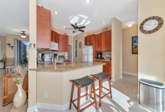 kitchen featuring kitchen peninsula, a kitchen bar, tasteful backsplash, light stone counters, and white appliances