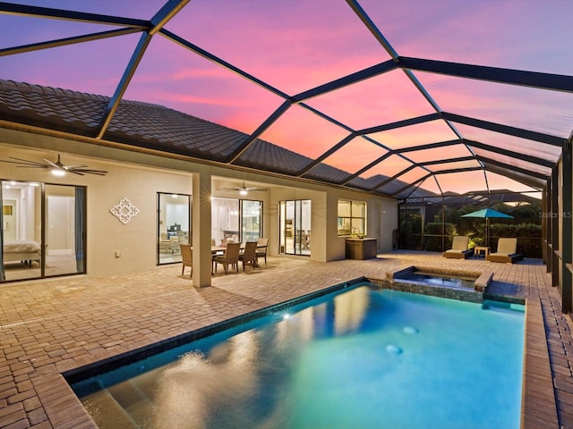 pool at dusk featuring glass enclosure, ceiling fan, a patio, and an in ground hot tub