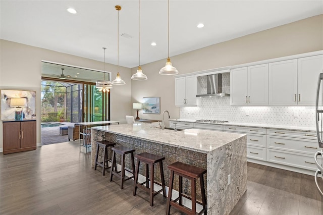 kitchen with white cabinets, a kitchen bar, wall chimney range hood, light stone counters, and a center island with sink