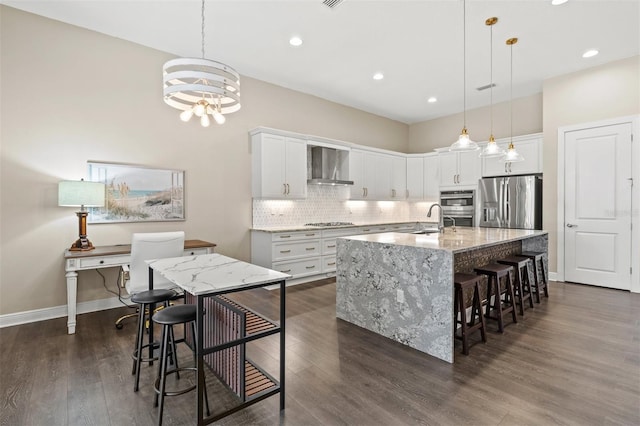 kitchen with white cabinetry, appliances with stainless steel finishes, a kitchen island with sink, wall chimney range hood, and sink