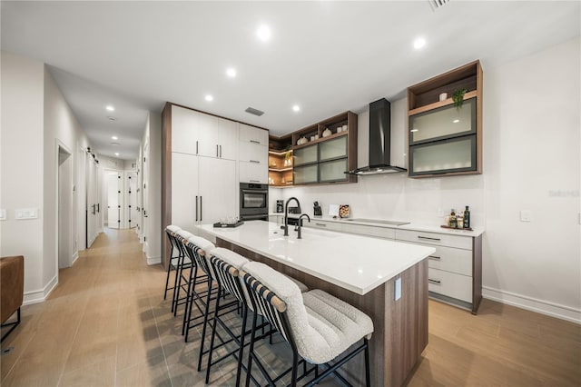 kitchen with wall chimney exhaust hood, black electric stovetop, oven, a kitchen island with sink, and a breakfast bar