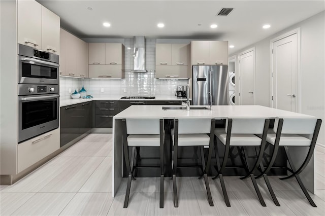 kitchen with appliances with stainless steel finishes, gray cabinetry, stacked washer / drying machine, a center island with sink, and wall chimney exhaust hood