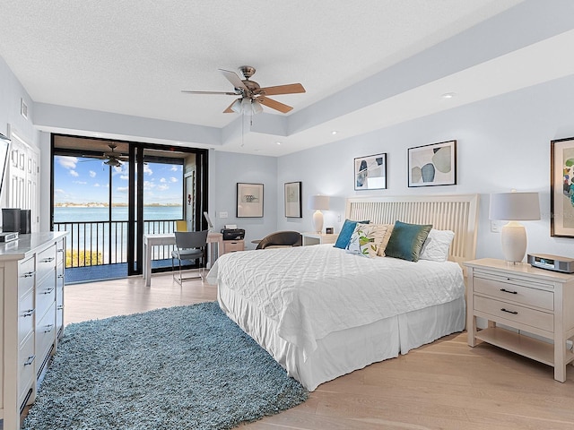 bedroom with ceiling fan, a tray ceiling, access to outside, a water view, and light wood-type flooring