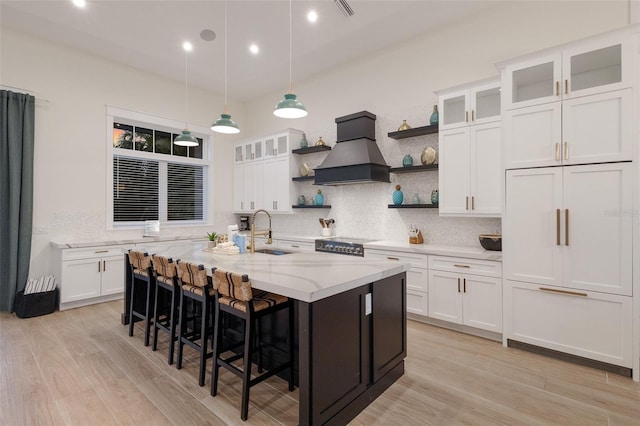 kitchen featuring light stone counters, custom range hood, pendant lighting, white cabinetry, and an island with sink