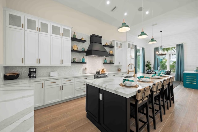 kitchen with light stone countertops, sink, an island with sink, white cabinets, and custom range hood