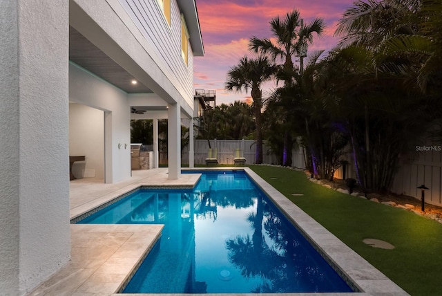 pool at dusk featuring a yard, an outdoor kitchen, a patio, and ceiling fan