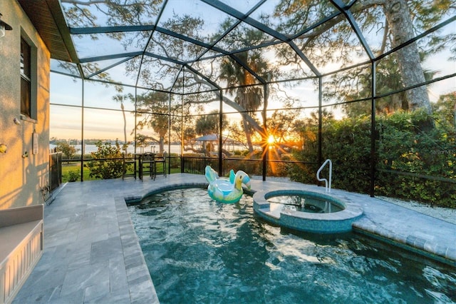 pool at dusk featuring glass enclosure, a pool with connected hot tub, and a patio area