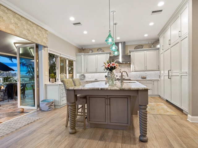 kitchen featuring pendant lighting, wall chimney range hood, sink, light stone countertops, and a center island with sink