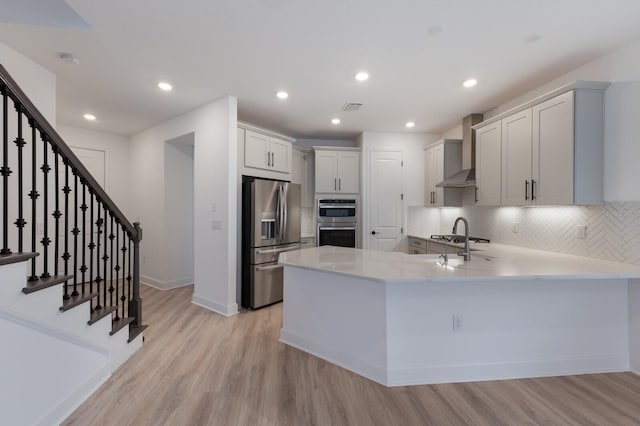 kitchen with white cabinetry, kitchen peninsula, stainless steel appliances, wall chimney range hood, and light hardwood / wood-style flooring