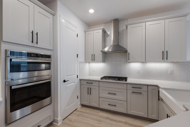 kitchen with backsplash, appliances with stainless steel finishes, light wood-type flooring, and wall chimney range hood