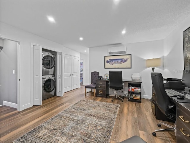 office with a wall mounted air conditioner, a textured ceiling, hardwood / wood-style flooring, and stacked washer / drying machine