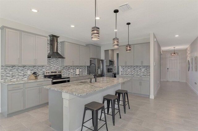 kitchen with stainless steel appliances, wall chimney range hood, a kitchen breakfast bar, light stone counters, and a kitchen island with sink