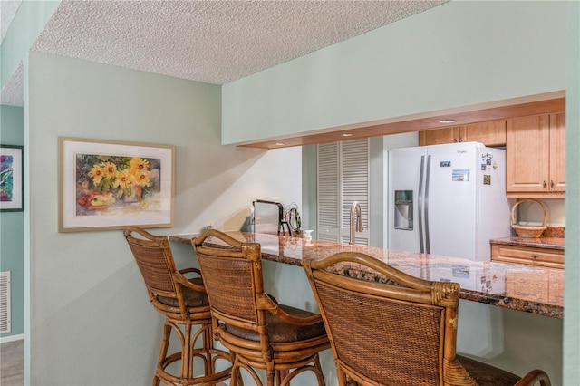 kitchen featuring a breakfast bar, a textured ceiling, white refrigerator with ice dispenser, light brown cabinets, and dark stone countertops