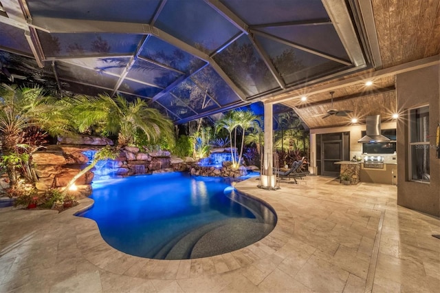 view of swimming pool featuring glass enclosure, ceiling fan, an outdoor kitchen, pool water feature, and a patio area