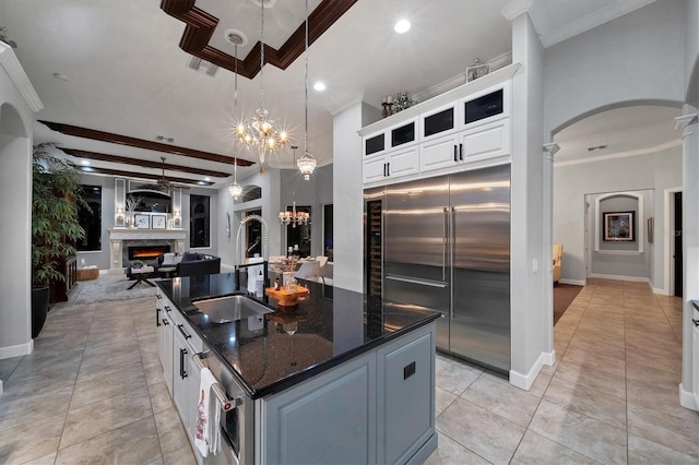 kitchen featuring a warm lit fireplace, a center island with sink, white cabinets, dark stone countertops, and stainless steel built in refrigerator