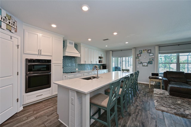 kitchen featuring black electric stovetop, custom range hood, double oven, a center island with sink, and white cabinets